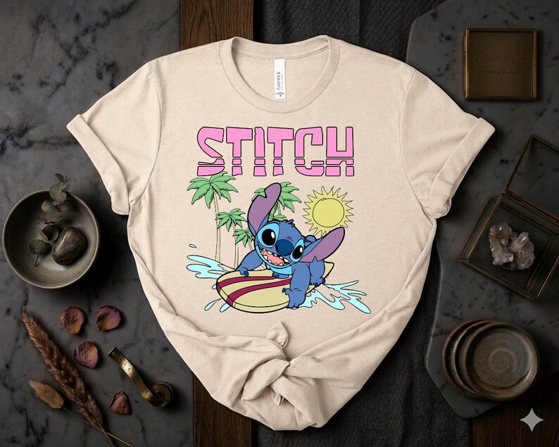 May include: Beige t-shirt featuring a graphic of Stitch surfing on a surfboard. The word "STITCH" is printed above the character in pink block letters. Palm trees and a yellow sun are also in the design.