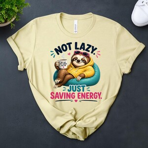 May include: A pale yellow t-shirt with a cartoon sloth design. The sloth is wearing sunglasses and a yellow hoodie, and is sitting on a blue beanbag. Text reads "NOT LAZY. JUST SAVING ENERGY."