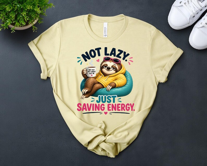 May include: A pale yellow t-shirt with a cartoon sloth design. The sloth is wearing sunglasses and a yellow hoodie, and is sitting on a blue beanbag. Text reads "NOT LAZY. JUST SAVING ENERGY."