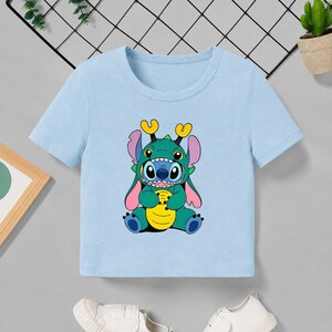 May include: A light blue t-shirt featuring a cartoon character in a green dragon costume. The character has blue skin, large eyes, and yellow horns. The shirt is short-sleeved and made of a soft material.