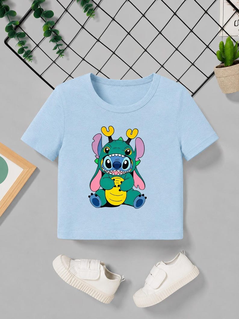 May include: A light blue t-shirt featuring a cartoon character in a green dragon costume. The character has blue skin, large eyes, and yellow horns. The shirt is short-sleeved and made of a soft material.