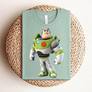 May include: A light green t-shirt featuring a colorful graphic of Buzz Lightyear from Toy Story. The character is depicted in his signature space ranger suit with green, white, and purple accents. The shirt is folded on a woven, circular surface.