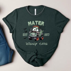 Może przedstawiać: Ciemnozielona koszulka z postacią z kresk&oacute;wki Mater z filmu Auta Disneya, w tematyce Halloween. Projekt zawiera napis "MATER EST 1977 DISNEY CARS" w upiornej czcionce, z nietoperzami i pajęczyną. Koszulka jest połączona z niebieskimi dżinsami.