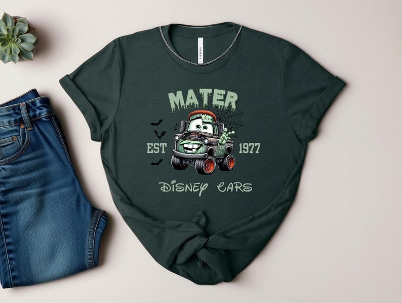 Może przedstawiać: Ciemnozielona koszulka z postacią z kresk&oacute;wki Mater z filmu Auta Disneya, w tematyce Halloween. Projekt zawiera napis "MATER EST 1977 DISNEY CARS" w upiornej czcionce, z nietoperzami i pajęczyną. Koszulka jest połączona z niebieskimi dżinsami.