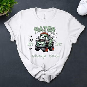 Może przedstawiać: Biała koszulka z halloweenowym motywem Matera z filmu Auta Disneya. Grafika zawiera tekst "MATER", "EST 1977" i "DISNEY CARS" w kolorze zielonym, wraz z nietoperzami i pajęczyną. Koszulka jest prezentowana na ciemnej powierzchni.