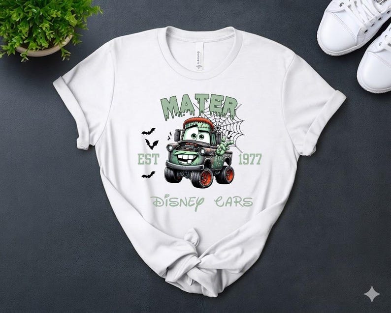 Może przedstawiać: Biała koszulka z halloweenowym motywem Matera z filmu Auta Disneya. Grafika zawiera tekst "MATER", "EST 1977" i "DISNEY CARS" w kolorze zielonym, wraz z nietoperzami i pajęczyną. Koszulka jest prezentowana na ciemnej powierzchni.
