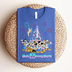 Może przedstawiać: Niebieska koszulka z kolorową grafiką postaci Disneya, zamkiem, fajerwerkami i napisem "Walt Disney World". Projekt zawiera Myszkę Miki i Minnie, Kaczora Donalda i Goofy'ego.