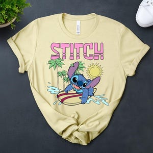 May include: A light yellow t-shirt featuring the word "STITCH" in pink block letters above a cartoon image of Stitch surfing. The graphic includes palm trees, a sun, and water splashes. The shirt is made of a soft material.