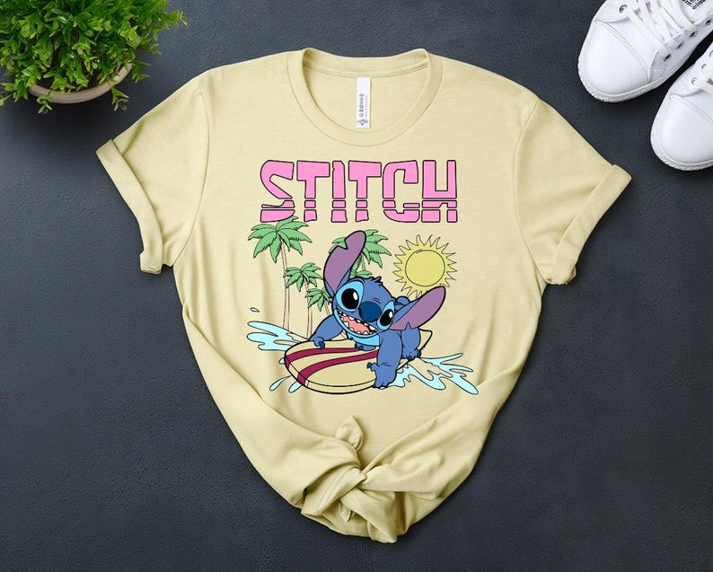 May include: A light yellow t-shirt featuring the word "STITCH" in pink block letters above a cartoon image of Stitch surfing. The graphic includes palm trees, a sun, and water splashes. The shirt is made of a soft material.
