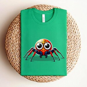Pu&ograve; includere: Una maglietta verde con un disegno di ragno dei cartoni animati. Il ragno ha una testa rotonda con grandi occhi, un corpo rosso e blu e zampe rosse e nere. La maglietta &egrave; appoggiata su una superficie circolare intrecciata.