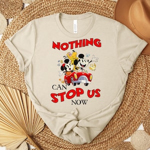 Puede incluir: Camiseta beige con una ilustraci&oacute;n de dibujos animados de Mickey y Minnie Mouse en un coche rojo, con el texto rojo "NOTHING CAN STOP US NOW". La camiseta tiene cuello redondo y mangas cortas.