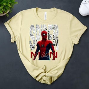 Peut inclure: Un t-shirt &agrave; manches courtes jaune clair avec un graphisme de Spider-Man en costume rouge et bleu. Le mot SPIDER est &eacute;crit en blanc sur un fond de cases de bande dessin&eacute;e en noir et blanc, et le mot MAN est imprim&eacute; en lettres rouges grasses.