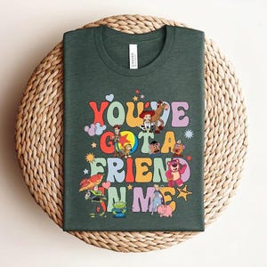May include: Dark green t-shirt with the text "You've Got a Friend in Me" in a retro font, accompanied by cartoon characters. The shirt is displayed on a woven, circular surface. The design is colourful and eye-catching.