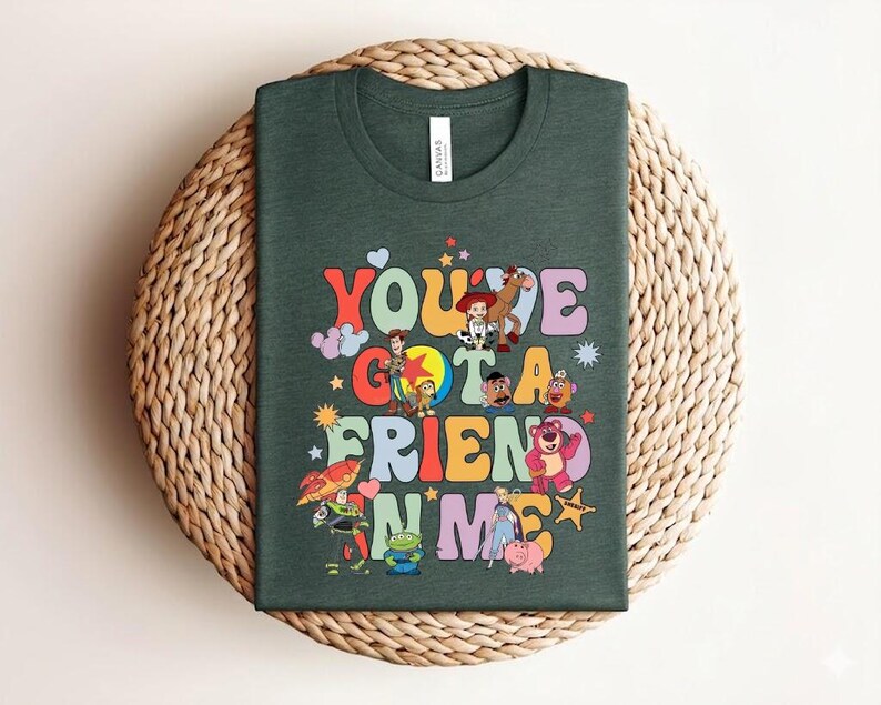 May include: Dark green t-shirt with the text "You've Got a Friend in Me" in a retro font, accompanied by cartoon characters. The shirt is displayed on a woven, circular surface. The design is colourful and eye-catching.
