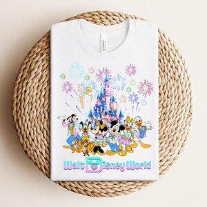Może przedstawiać: Biała koszulka z kolorową grafiką postaci Disneya, zamku i fajerwerk&oacute;w. Tekst "Walt Disney World" jest wyświetlany na dole. Koszulka jest złożona na tkanej, okrągłej powierzchni.