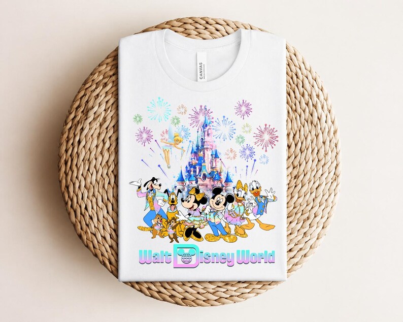 Może przedstawiać: Biała koszulka z kolorową grafiką postaci Disneya, zamku i fajerwerk&oacute;w. Tekst "Walt Disney World" jest wyświetlany na dole. Koszulka jest złożona na tkanej, okrągłej powierzchni.