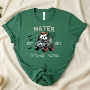 Może przedstawiać: Zielona koszulka z postacią z kresk&oacute;wki Mater z Disney Cars, w tematyce Halloween. Projekt zawiera napis "Mater Est 1977 Disney Cars" z nietoperzami i pajęczyną.