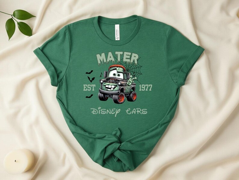 Może przedstawiać: Zielona koszulka z postacią z kresk&oacute;wki Mater z Disney Cars, w tematyce Halloween. Projekt zawiera napis "Mater Est 1977 Disney Cars" z nietoperzami i pajęczyną.