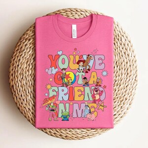 May include: A pink t-shirt with the text "You've Got a Friend in Me" in colourful, bubble-style letters. The shirt features cartoon characters such as Woody and Buzz Lightyear, in a playful design.