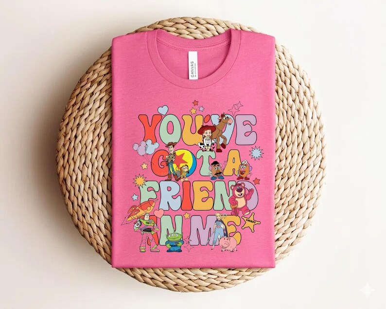May include: A pink t-shirt with the text "You've Got a Friend in Me" in colourful, bubble-style letters. The shirt features cartoon characters such as Woody and Buzz Lightyear, in a playful design.