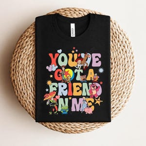 May include: Black t-shirt with the phrase "You've Got a Friend in Me" in colourful, retro lettering. The design features cartoon characters from a popular animated film. A fun, playful design for fans of the film.