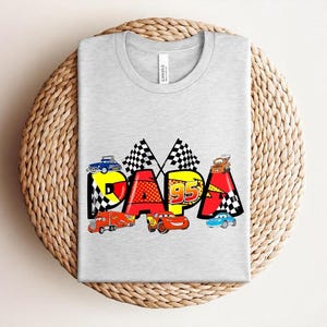 Puede incluir: Camiseta gris claro con la palabra "PAPA" en un dise&ntilde;o colorido con coches de carreras y banderas a cuadros. El dise&ntilde;o incluye el n&uacute;mero "95" e ilustraciones de coches, ideal para un aficionado a los coches.