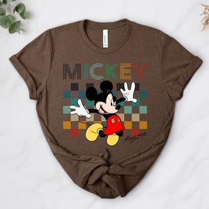 May include: Brown t-shirt featuring Mickey Mouse with the word "MICKEY" in colorful block letters. Mickey is depicted with his arms raised, wearing white gloves, red shorts, and yellow shoes. The background has a checkered pattern.