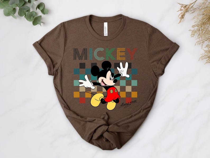 May include: Brown t-shirt featuring Mickey Mouse with the word "MICKEY" in colorful block letters. Mickey is depicted with his arms raised, wearing white gloves, red shorts, and yellow shoes. The background has a checkered pattern.
