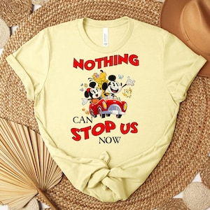 Puede incluir: Camiseta amarillo claro con Mickey y Minnie Mouse en un coche rojo y el texto "NOTHING CAN STOP US NOW" en rojo. La camiseta tiene cuello redondo y mangas cortas.