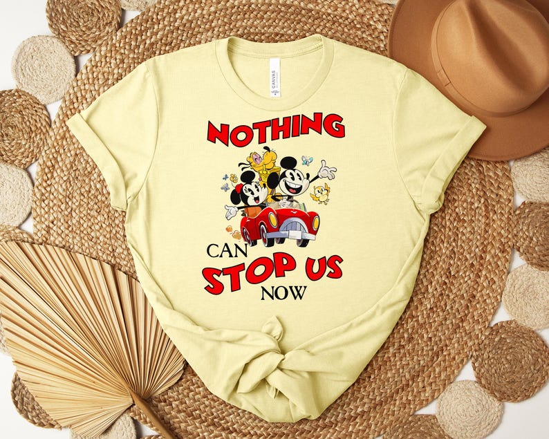 Puede incluir: Camiseta amarillo claro con Mickey y Minnie Mouse en un coche rojo y el texto "NOTHING CAN STOP US NOW" en rojo. La camiseta tiene cuello redondo y mangas cortas.