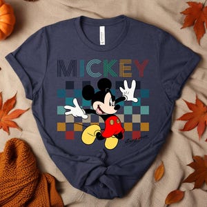 May include: Navy blue t-shirt featuring Mickey Mouse with the word "MICKEY" in colorful block letters. Mickey is depicted in his classic pose, with white gloves and yellow shoes. The shirt has a checkered background.