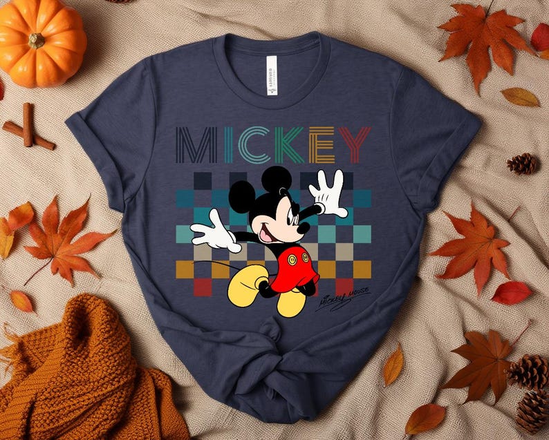 May include: Navy blue t-shirt featuring Mickey Mouse with the word "MICKEY" in colorful block letters. Mickey is depicted in his classic pose, with white gloves and yellow shoes. The shirt has a checkered background.