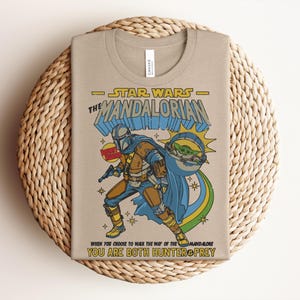 K&ouml;nnte beinhalten: Beiges T-Shirt mit einem farbenfrohen Grafikdesign von The Mandalorian aus Star Wars. Das Design enth&auml;lt den Text "STAR WARS THE MANDALORIAN" und das Zitat "WHEN YOU CHOOSE TO WALK THE WAY OF THE MANDALORE YOU ARE BOTH HUNTER & PREY."