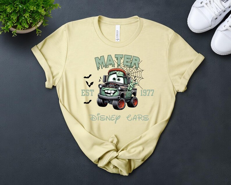 Może przedstawiać: Jasnoż&oacute;łta koszulka z motywem zombie Mater z filmu Auta Disneya. Projekt zawiera tekst "MATER", "EST 1977" i "Disney Cars" w kolorze zielonym, z grafiką nietoperzy i pajęczyny. Odzież codzienna.