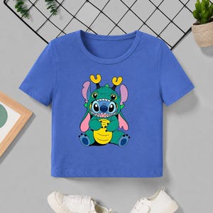 May include: A blue short-sleeved t-shirt featuring a cartoon character in a green and blue costume. The character has large eyes, yellow horns, and pink ears. The graphic is centered on the shirt.