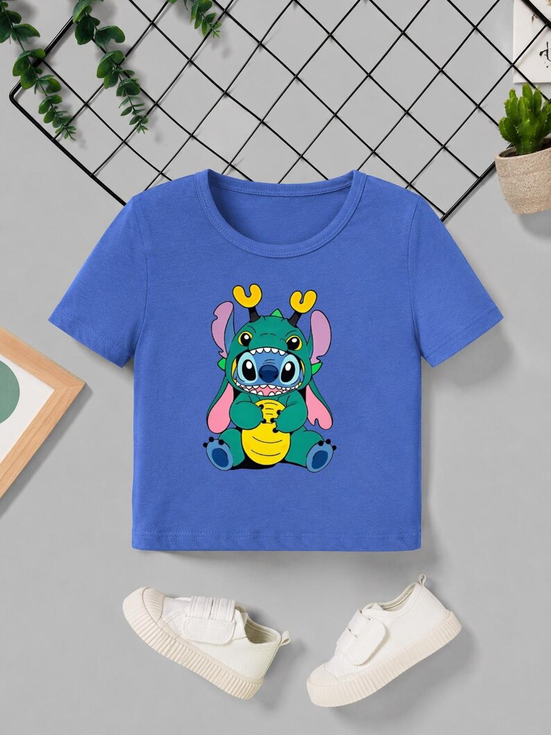 May include: A blue short-sleeved t-shirt featuring a cartoon character in a green and blue costume. The character has large eyes, yellow horns, and pink ears. The graphic is centered on the shirt.