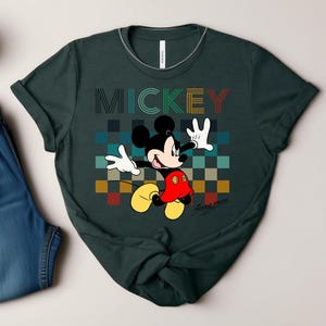 May include: Dark green t-shirt featuring a colorful graphic of Mickey Mouse. The word "MICKEY" is printed above the character in a retro font. Mickey Mouse is depicted with his signature yellow shoes, red shorts, and white gloves.
