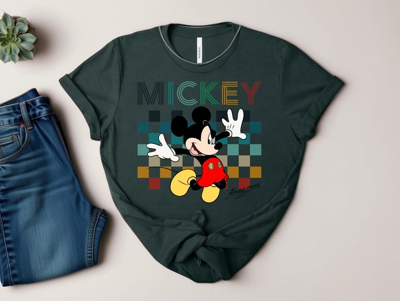 May include: Dark green t-shirt featuring a colorful graphic of Mickey Mouse. The word "MICKEY" is printed above the character in a retro font. Mickey Mouse is depicted with his signature yellow shoes, red shorts, and white gloves.