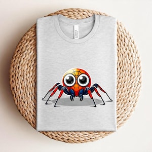Pu&ograve; includere: T-shirt grigio chiaro con un design di ragno dei cartoni animati colorato. Il ragno ha un corpo rotondo con sfumature gialle, arancioni e rosse, grandi occhi e zampe rosse e nere. La maglietta &egrave; piegata.