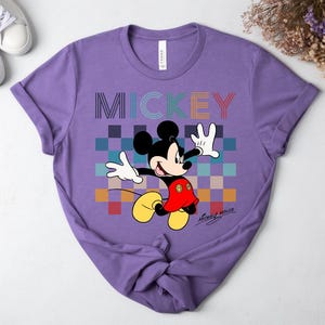 May include: A purple t-shirt featuring a colorful graphic of Mickey Mouse. The word "MICKEY" is printed across the top in a rainbow-colored font. Mickey Mouse is depicted in his classic pose, with a red shirt, yellow shoes, and white gloves.