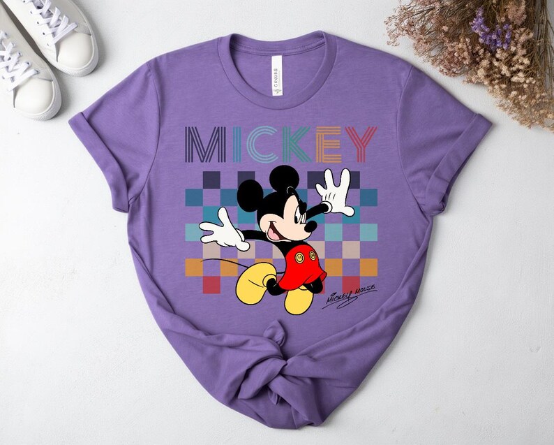 May include: A purple t-shirt featuring a colorful graphic of Mickey Mouse. The word "MICKEY" is printed across the top in a rainbow-colored font. Mickey Mouse is depicted in his classic pose, with a red shirt, yellow shoes, and white gloves.