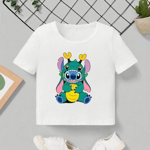 May include: White short-sleeved t-shirt featuring a colorful cartoon character design. The graphic depicts a blue and green creature with large ears and a yellow belly. The shirt is paired with white sneakers.