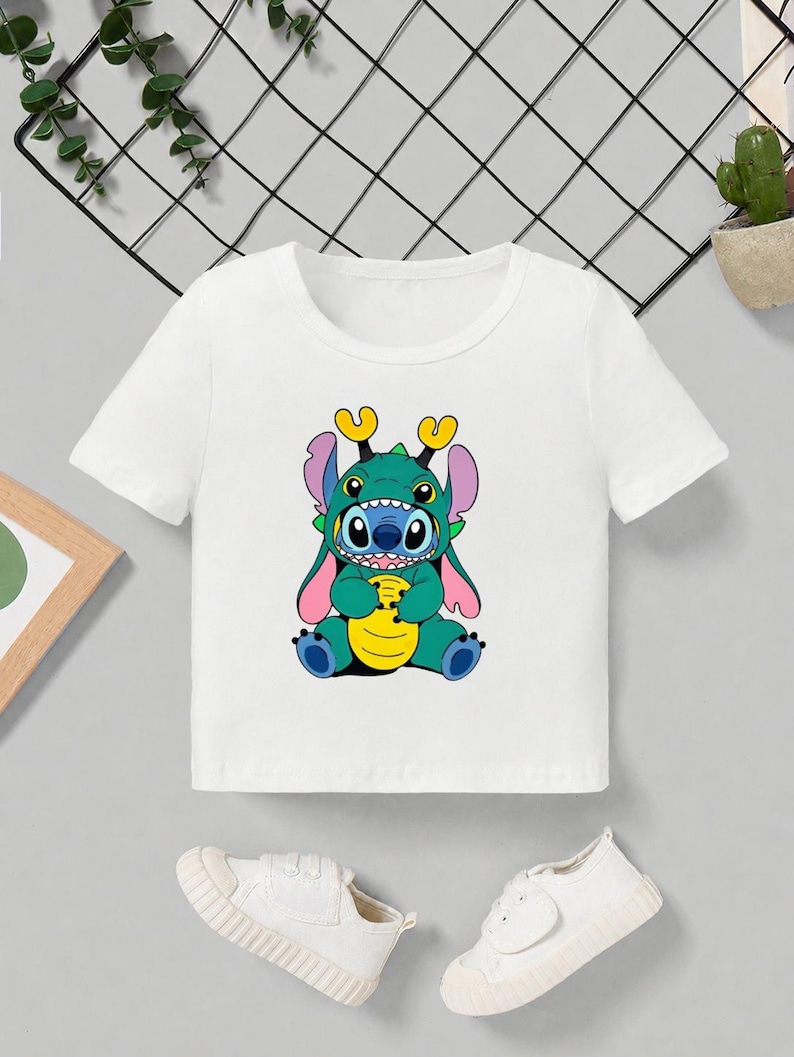 May include: White short-sleeved t-shirt featuring a colorful cartoon character design. The graphic depicts a blue and green creature with large ears and a yellow belly. The shirt is paired with white sneakers.