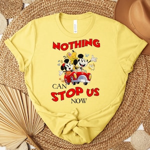 Puede incluir: Camiseta amarilla con un gr&aacute;fico de dibujos animados de Mickey y Minnie Mouse en un coche rojo, con el texto "NOTHING CAN STOP US NOW" en rojo. El dise&ntilde;o incluye otros personajes de dibujos animados y mariposas.