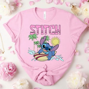 May include: Pink t-shirt featuring the word "STITCH" in pink and a cartoon image of Stitch surfing. The design includes palm trees, a yellow sun, and blue water. The shirt is a crew neck style.