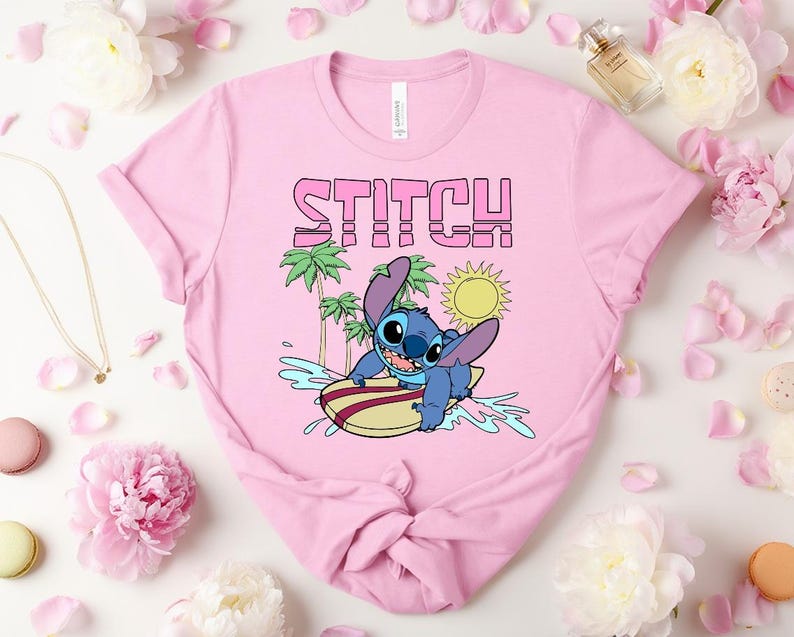 May include: Pink t-shirt featuring the word "STITCH" in pink and a cartoon image of Stitch surfing. The design includes palm trees, a yellow sun, and blue water. The shirt is a crew neck style.