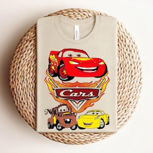 May include: Beige t-shirt featuring a colorful graphic of characters from the animated movie "Cars." The design includes a red race car, a brown tow truck, and a yellow car, with the word "Cars" prominently displayed.