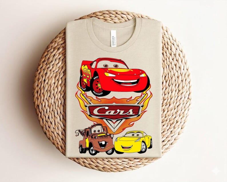 May include: Beige t-shirt featuring a colorful graphic of characters from the animated movie "Cars." The design includes a red race car, a brown tow truck, and a yellow car, with the word "Cars" prominently displayed.