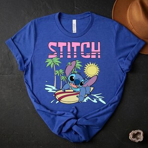 May include: Blue t-shirt featuring the word "STITCH" in pink block letters above a cartoon image of Stitch surfing. The graphic includes a yellow sun, palm trees, and ocean waves. The shirt is made of a soft material.
