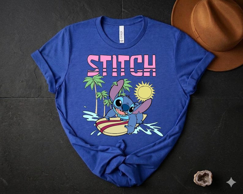 May include: Blue t-shirt featuring the word "STITCH" in pink block letters above a cartoon image of Stitch surfing. The graphic includes a yellow sun, palm trees, and ocean waves. The shirt is made of a soft material.